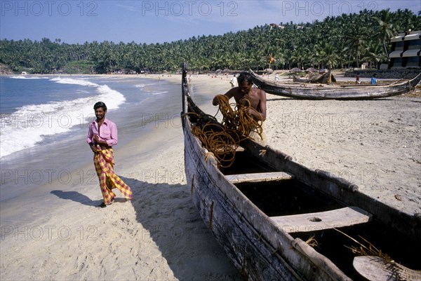 INDIA, Kerala, Kovalam, Fisherman tending to his boat moored on the sandy beach with passing man in sarong