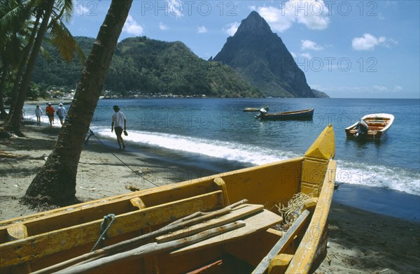 WEST INDIES, St Lucia, Soufriere, People walking past fishing boats on beach with the Pitons in the distance