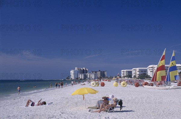 USA, Florida, St.Petersburg, Woman reading under a multicoloured parasol on a sandy beach with hotels in the distance