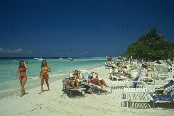 WEST INDIES, Jamaica, Negril, Female tourists beach sunbahing on sun loungers by the water with two women in swimsuits walking past