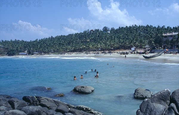 INDIA, Kerala, Kovalam, View from rocky outcrop over bay toward beach surrounded by palms with group of bathers in the sea