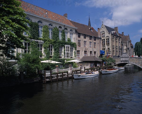 BELGIUM, West Flanders, Bruges, "Boats dock on the canal near a bridge, next to an ivy covered building with canopies."
