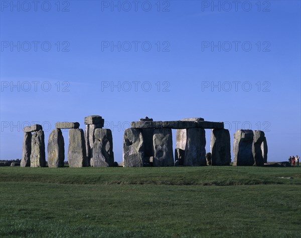 ENGLAND, Wiltshire, Stonehenge, View across the grass on Salisbury Plain toward standing stones with visitors at base