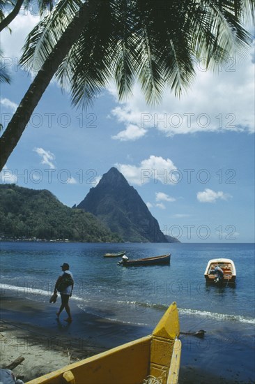 WEST INDIES, St Lucia, Soufriere, Man walking along beach with boats moored offshore and the Pitons behind.  Prow of boat and palm in foreground.