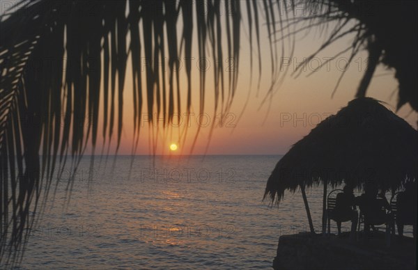 WEST INDIES, Jamaica, Negril, Ricks Cafe at sunset through coconut palm tree with tourist sitting under thatched sun shade