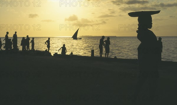 TANZANIA , Zanzibar, Zanzibar, People on the waterfront silhouetted against yellow sunset with dhow at sea in the distance