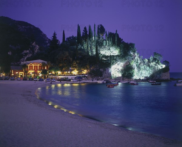 GREECE, Epirus Region, Parga, View along Krioneri Beach to restaurant and rocky headland illuminated at night