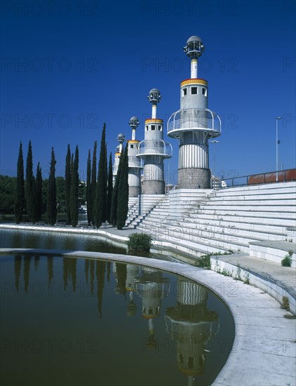 SPAIN, Catalonia, Barcelona, Parc Espanya Industrial.  Line of towers with balconies encircling their walls and spruce trees reflected in circular pond in the foreground.