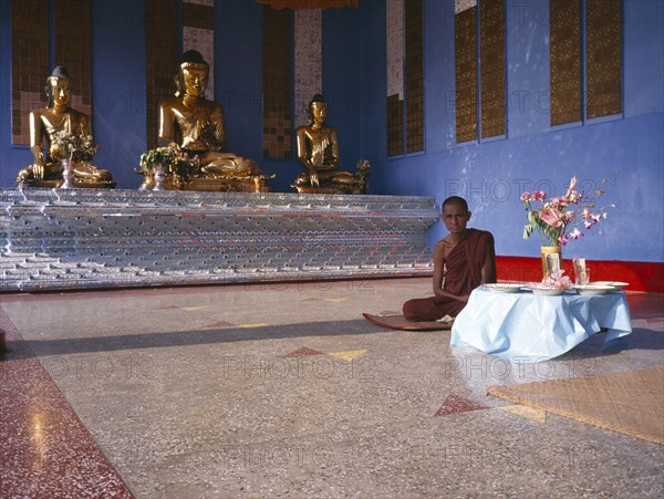 MYANMAR, Yangon, "Shwedagon Pagoda interior.  Monk sitting beside a raised altar with three seated, golden Buddha figures.   "
