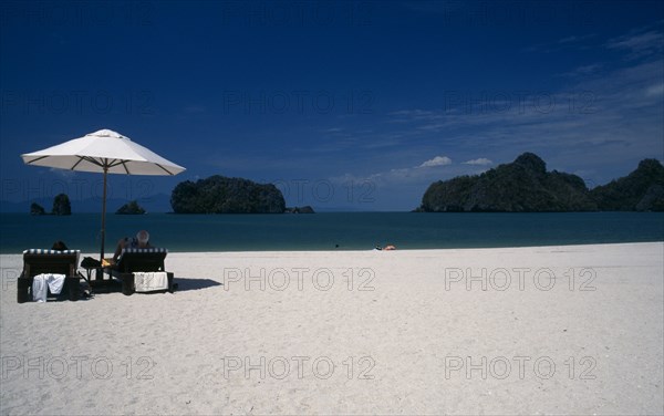 MALAYSIA, Kedah, Langkawi, Pantai Rhu beach with tourists sitting under an umbrella looking out to the offshore islands