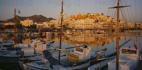 GREECE, Cyclades Islands, Naxos, Hora.  View over moored boats and harbour towards town reflected in the water in evening light.