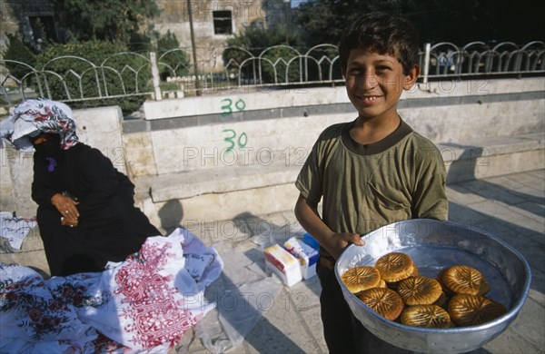 SYRIA, Central, Hama, "Young boy selling sweetmeats in the market, woman in traditional black dress and veil sitting on pavement behind."