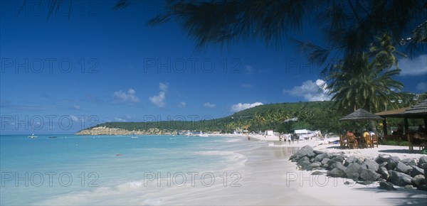 WEST INDIES, Antigua, Dickenson Bay, View along beach towards tree covered headland with people walking along shoreline and sitting under thatched sun umbrellas nearby.