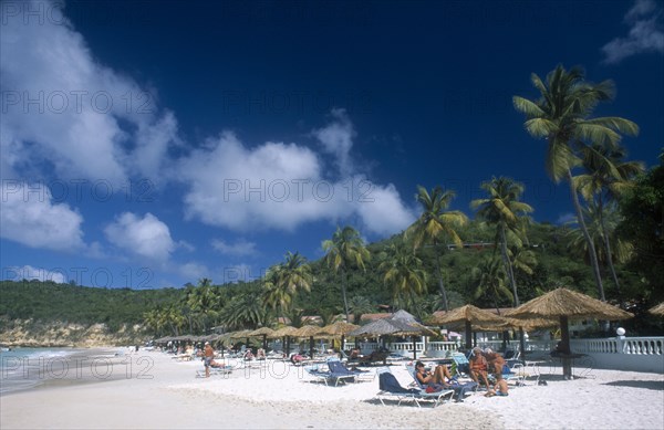 WEST INDIES, Antigua, Dickenson Bay, View along sandy beach lined with palm trees and thatched sun umbrellas with people sunbathing on loungers.