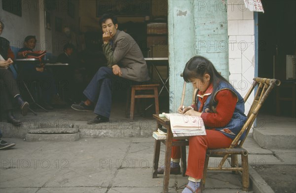 CHINA, Sichuan, Leshan, Young girl doing homework at small table outside house watched by adults in open doorway behind with one smoking.