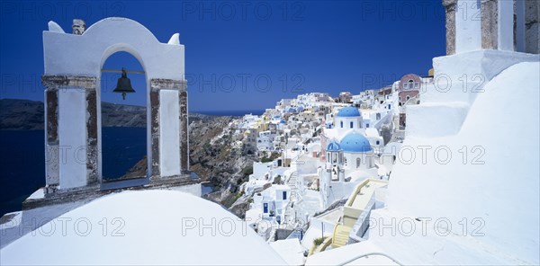 GREECE, Cyclades Islands, Santorini, Oia.  White painted hill town with bell in foreground hanging from arch part framing view over sea to coastline.