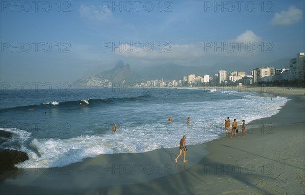 BRAZIL, Rio State, Rio de Janeiro, Ipanema Beach with bathers standing at the waters edge in early morning light with misty mountains in the distance.
