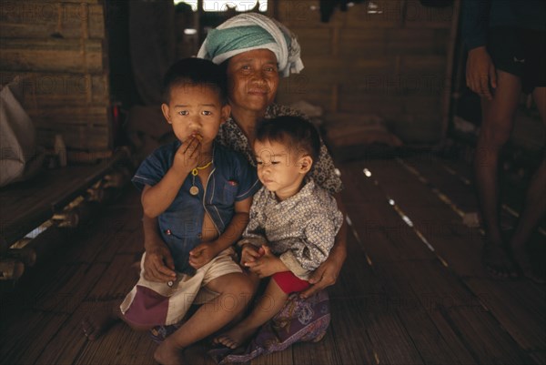 THAILAND, North, Mae Sai Area, Karen Refugee mother sitting with her two children on the floor of a hut