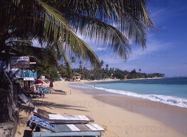 SRI LANKA, Unawatuna, Narrow sandy beach lined with vegetation and overhanging palm trees with hotel restaurant and wooden sun loungers.