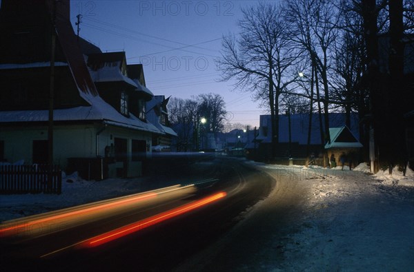 POLAND, Zakopane, Chocholow, Streaked red car lights in motion blur on road through snowy village at dusk