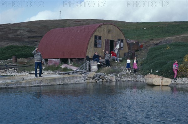FALKLAND ISLANDS, West Point Island, View across water towards loading jetty with a family near building