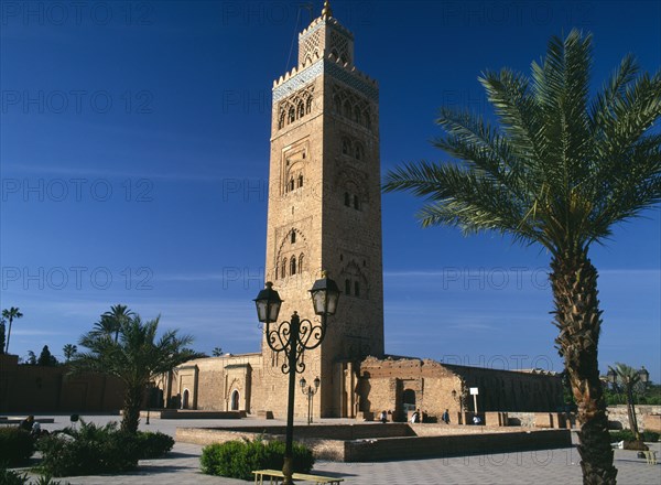 MOROCCO, Marrakech, Koutoubia Mosque tower seen from pavement with blue sky behind and palm trees in foreground
