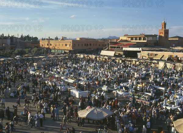 MOROCCO, Marrakech, Djemaa El Fina Square. View over busy market square with crowds of people at stalls and buildings behind.