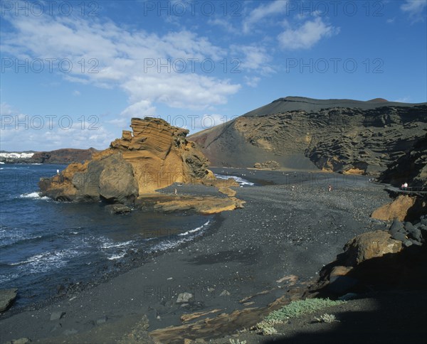 SPAIN, Canary Islands, Lanzarote, El Golfo.  Black volcanic sand beach and coastline with a few people sunbathing or walking along shore.