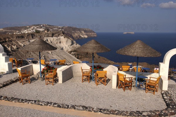GREECE, Cyclades, Santorini, Terrace with tables and chairs under thatch umbrellas overlooking coastal cliffs