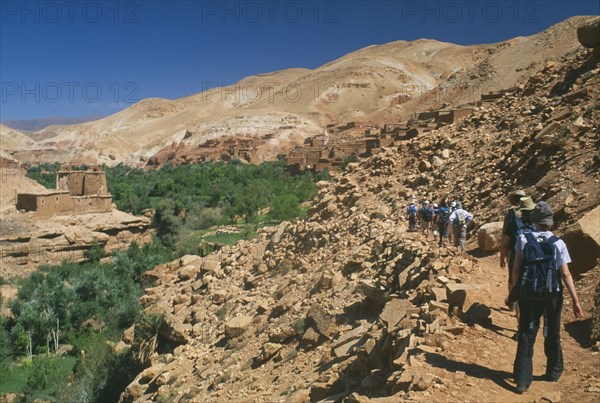 MOROCCO, High Atlas Mountains, General, "Tourist group walking along narrow, rocky path towards flat roofed housing set into steep hillside with fertile valley gorge on left."