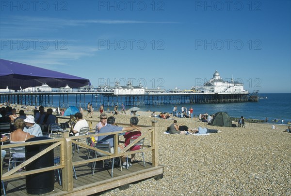 ENGLAND, East Sussex, Eastbourne, Beach cafe with people sat on chairs under parasols infront of busy shingle beach and pier