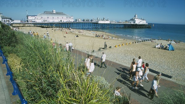 ENGLAND, East Sussex, Eastbourne, View from promenade towards shingle beach and pier with people walking on path.