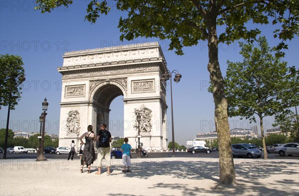 FRANCE, Ile de France, Paris, Tourist family standing in the shade of a tree in Place de Charles de Gaulle looking at the Arc de Triomphe with traffic passing around it