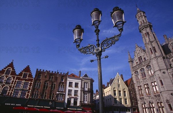 BELGIUM, West Flanders, Bruges, The Markt (Market Place).  Line of cafe awnings and red umbrellas of outside tables with street lamp in the foreground.