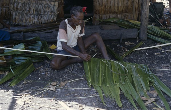 PACIFIC ISLANDS, Melanesia, Solomon Islands, "Malaita Province, Lau Lagoon, Foueda Island.  Traditional building methods, man stitching lengths of palm leaves to narrow strip of wood to use in house construction."