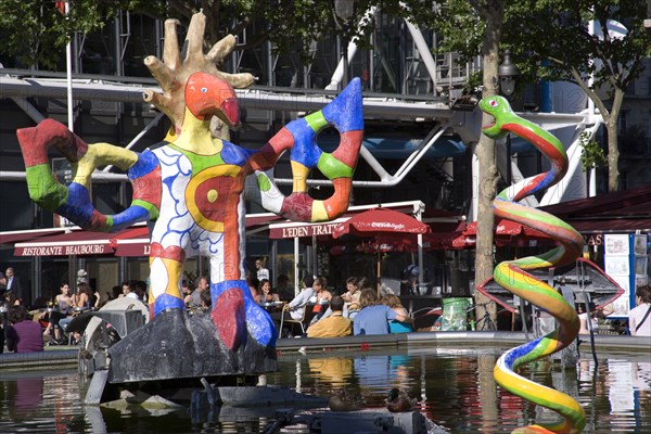FRANCE, Ile de France, Paris, People sitting at tables under umbrellas beside the colourful contemporary fountains by Niki de Saint Phalle and Jean Tinguely in Place Igor Stravinsky beside the Pompidou Centre in Beauborg Les Halles