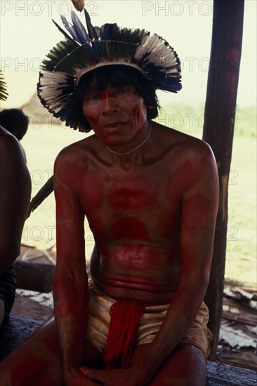 BRAZIL, Mato Grosso, Indigenous Park of the Xingu, Portrait of Panara elder Kokridi  with face and body painted red for dance wearing crown or head-dress of feathers. Formally known as Kreen-Akrore  Krenhakarore  Krenakore  Krenakarore  Amazon American Brasil Brazilian Indegent Kreen Akore Latin America Latino South America Immature One individual Solo Lone Solitary