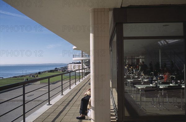 ENGLAND, East Sussex, Bexhill-on-Sea, De La Warr Pavilion. View along the sun terrace next to the restaurant with people sitting on benches behind a pillar looking out to the sea.
