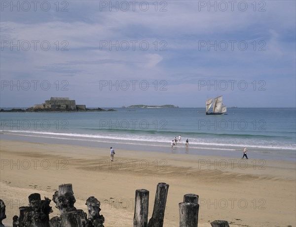 FRANCE, Brittany, St Malo, Le Sillon Beach and Fort National. People walking along golden sand with a sail boat on seen on water
