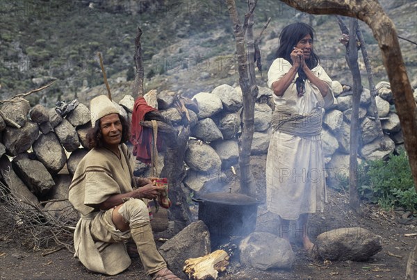 COLOMBIA, Sierra Nevada de Santa Marta, Ika, Ika brother and sister in traditional woven wool&cotton mantas cloaks  helmet of woven fique cactus fibre and wool waist belts  cook a potato and mutton sancocho - stew  outside their home using aluminium cooking pot placed on rounded boulders over wood fire. Arhuaco Aruaco indigenous tribe American Colombian Colombia Hispanic Indegent Latin America Latino South America  Arhuaco Aruaco indigenous tribe American Colombian Columbia Hispanic Indegent Latin America Latino South America 2 Aluminum Classic Classical Fiber Historical Older Two