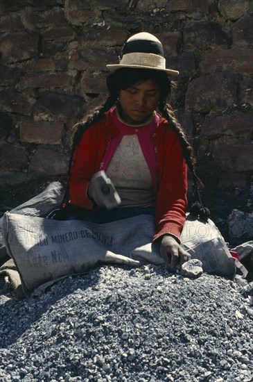 BOLIVIA, Chukiuta, Woman sitting in a Banco Minero de Bolivia sack breaking rocks into small pieces with a hammer at a mine. Near Sucre