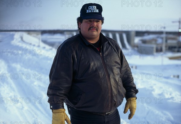 CANADA, Quebec, James Bay, Man named Larry who is a Cree indigenous activist wearing a Cree Nation baseball cap standing in the snow in front of Hydro Quebec’s giant new dam project which has ruined Cree fishing downstream of the dam