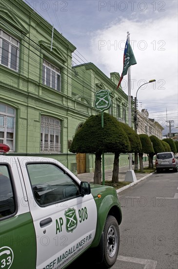 Punta Arenas, Magellan and Antarctic Region, Chile. Green painted Police Headquarters off the Plaza Munoz Gamero with a green and white police car parked outside and the crossed rifles logo of the Carabineros de Chile visible on the car sign and flag. Manicured green trees on the sidewalk. Chile Chilean Magallanes South America American Hispanic Santiago City Cityscape Urban Magallanes Punta Arenas Police HQ Headquarters Station Plaza Munoz Gamero Green Carabibneros Car Jeep Vehicle Automobile Automotive Cars Color Latin America Latino Motorcar Signs Display Posted Signage South America Southern