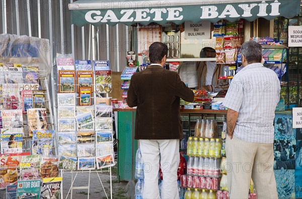 Istanbul, Turkey. Sultanahmet. Customers at newspaper kiosk with magazine stand and display of maps postcards snacks and soft drinks. Turkey Turkish Istanbul Constantinople Stamboul Stambul City Europe European Asia Asian East West Urban Destination Travel Tourism Sultanahmet Market Stall Selling Vendor News Newstand Newspaper Newspapers Kiosk Booth Customers Buying men Destination Destinations Male Man Guy Middle East South Eastern Europe Turkiye Western Asia