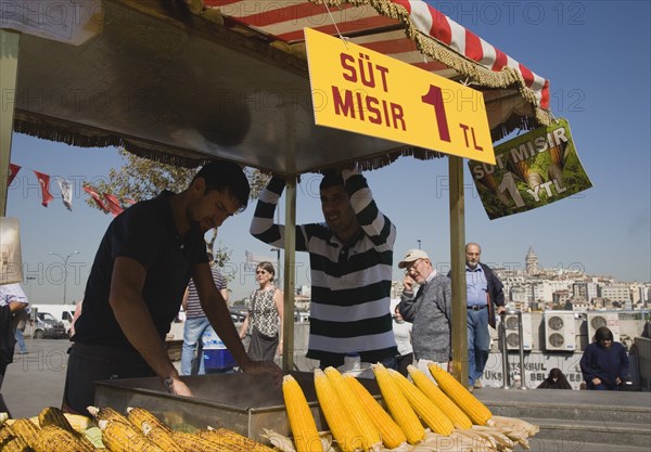 Istanbul, Turkey. Sultanahmet. Street stall selling freshly grilled corn on the cob outside The New Mosque or Yeni Camii with Galata Bridge behind. Turkey Turkish Istanbul Constantinople Stamboul Stambul City Europe European Asia Asian East West Urban Destination Travel Tourism Sultanahmet Street Stall Vendor Food Grilled Corn Cob Fast Market Color Destination Destinations Middle East South Eastern Europe Turkiye Western Asia