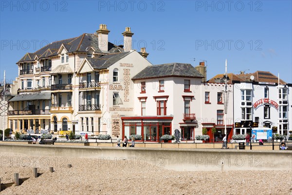 England, West Sussex, Bognor Regis, The Royal Hotel Bar and Restaurant and other buildings on the Esplanade with people walking on the promenade behind the sea wall defences on the pebble shingle beach.