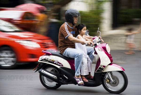 Father with young daughter on motorcycle pass brightly coloured taxis.